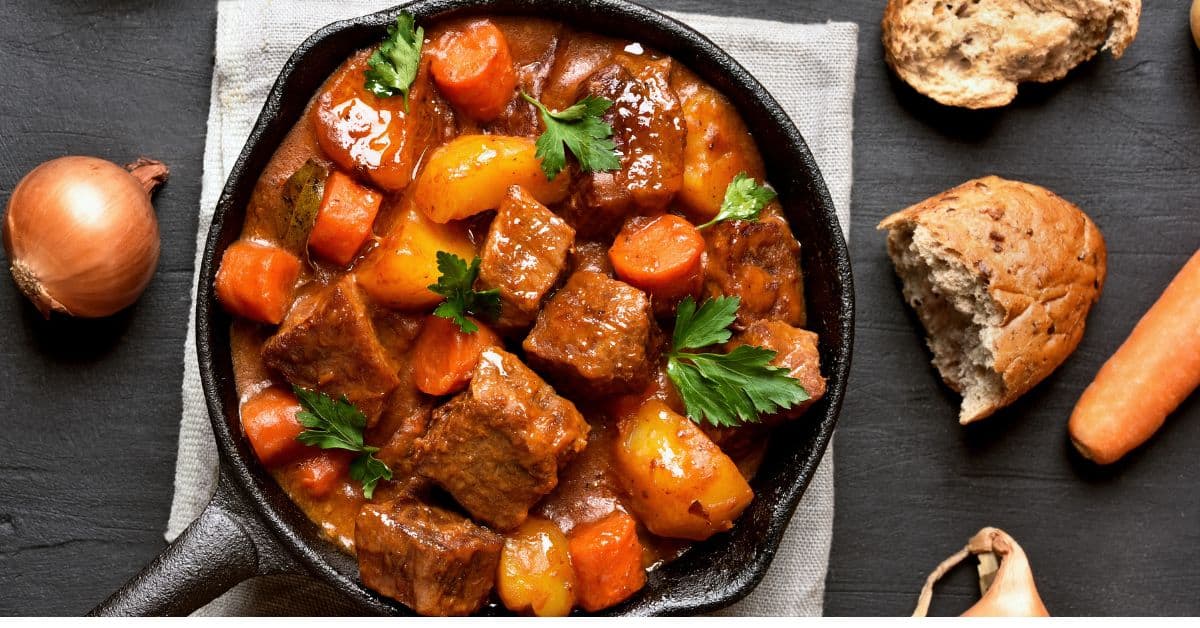 Beef Stew in cast iron skillet with bread and onions around the boarder. The stew has large chunks of beef with halved potatoes and carrots. Fresh green Parsley sprinkled on top